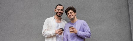 Positive and young same sex couple holding takeaway coffee in paper cups while standing near building on urban street, banner 