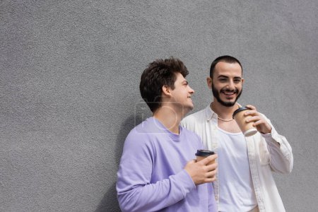 Happy bearded homosexual man holding coffee to go near boyfriend in purple sweatshirt and braces while standing near building on urban street 