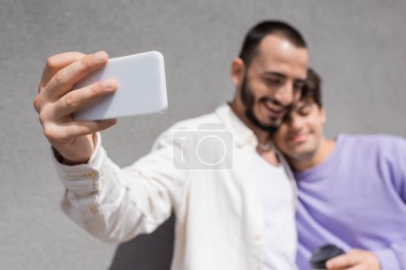 Téléchargez les photos : Couple de même sexe flou en vêtements décontractés prenant selfie sur smartphone ensemble tout en se tenant près du mur du bâtiment sur la rue urbaine le jour - en image libre de droit