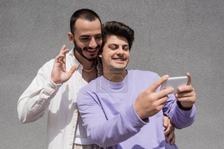 Sonriendo pareja gay en ropa casual teniendo videollamada en un teléfono inteligente borroso mientras están de pie juntos cerca del edificio en la calle urbana durante el día  