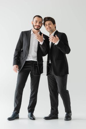 Full length of positive same sex grooms in elegant classic suits showing golden wedding rings at camera while standing on grey background 