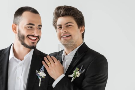 Portrait of cheerful gay groom in classic suit with braces and floral boutonniere hugging cheerful bearded boyfriend during wedding celebration isolated on grey  