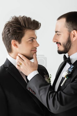 Portrait of carefree homosexual groom in formal wear with floral boutonniere touching chin of young boyfriend during wedding celebration isolated on grey  