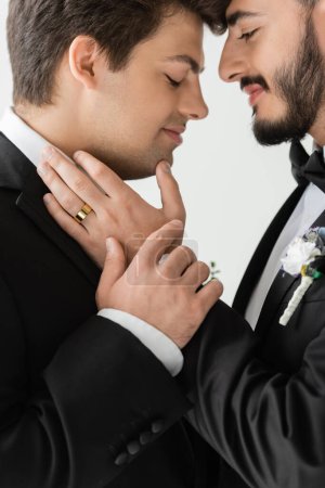 Foto de Vista lateral de los novios jóvenes del mismo sexo en trajes clásicos con boutonnieres tocándose entre sí durante la celebración de la boda y mientras están de pie aislados en gris - Imagen libre de derechos