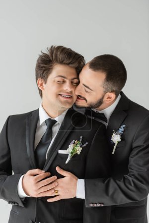 Young and bearded gay groom in elegant suit with boutonniere hugging and kissing smiling boyfriend with closed eyes and braces while celebrating wedding isolated on grey  
