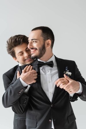 Lächelnder homosexueller Bräutigam mit geschlossenen Augen umarmt eleganten Freund im klassischen Anzug mit Boutonniere während Hochzeitsfeier isoliert auf grau  