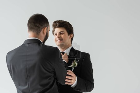 Homosexuelle Mann in formeller Kleidung Anpassung Krawatte von positiven und brünetten Bräutigam mit boutonniere und Zahnspange während der Hochzeitsfeier isoliert auf grau