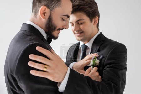 Positive and bearded gay groom adjusting floral boutonniere on elegant suit of blurred boyfriend in braces during wedding ceremony isolated on grey  