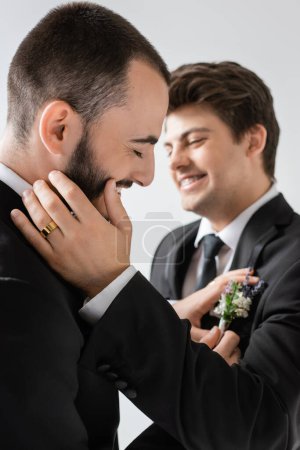 Cheerful bearded gay groom adjusting blurred floral boutonniere on suit of blurred smiling young boyfriend in braces during wedding celebration isolated on grey  