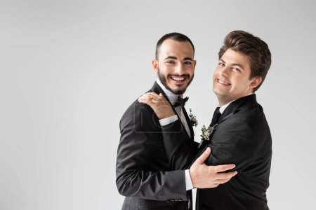 Positive gay man in formal wear with boutonniere hugging young bearded boyfriend in braces and looking at camera during wedding while standing together isolated on grey 