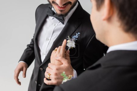 Ausgeschnittene Ansicht des verschwommenen homosexuellen Bräutigams Anpassung boutonniere auf Anzug des lächelnden Freundes während der Hochzeitsfeier isoliert auf grau 