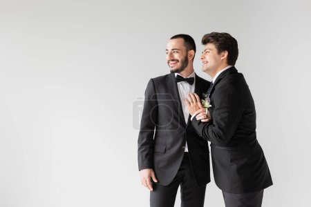 Junger homosexueller Bräutigam im eleganten Anzug mit floralem Boutonniere berührt fröhlichen Freund in Hosenträgern und schaut während der Hochzeitsfeier isoliert auf grau 