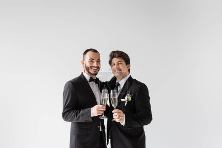 Pareja sonriente y joven del mismo sexo en trajes elegantes con boutonniere floral abrazando y sosteniendo copas de champán durante la ceremonia de boda aislada en gris 