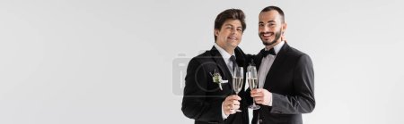 Positive same sex couple in elegant suits holding glasses of champagne and looking at camera during wedding ceremony isolated on grey, banner 
