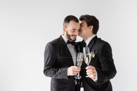 Young homosexual man in suit with boutonniere whispering in ear of smiling boyfriend with glass of champagne during wedding ceremony isolated on grey 