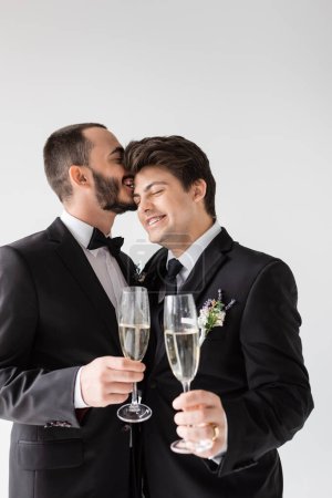 Smiling gay groom in suit with boutonniere whispering to young boyfriend in braces  and holding glass of champagne during wedding ceremony isolated on grey 