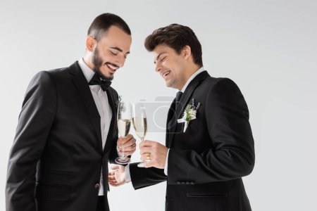 Unbekümmert und bärtig prostet homosexueller Bräutigam Champagnerglas mit elegantem Freund in Hosenträgern mit Boutonniere im Anzug während der Hochzeitszeremonie isoliert auf grau an 