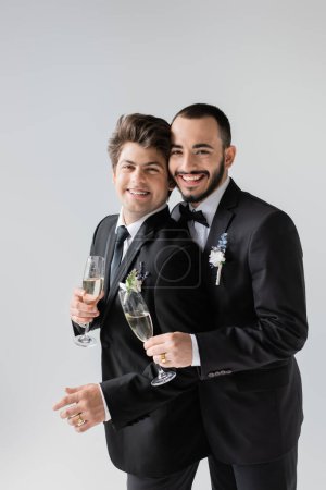 Retrato de pareja positiva del mismo sexo en elegantes trajes clásicos sosteniendo copas de champán mientras están de pie juntos durante la celebración de la boda aislados en gris 
