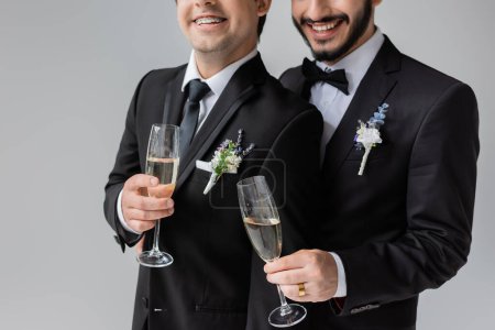 Foto de Vista recortada de novios homosexuales sonrientes con atuendo clásico con boutonnieres florales sosteniendo copas de champán mientras celebran la boda aislada en gris - Imagen libre de derechos
