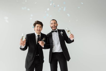 Excited same sex grooms in classic suits with boutonnieres holding hands and glasses of champagne while standing under falling confetti during wedding celebration on grey background