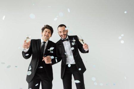 Aufgeregte homosexuelle Bräutigame in eleganter offizieller Kleidung halten Händchen und Sektgläser, während sie während der Hochzeit unter fallendem Konfetti auf grauem Hintergrund stehen