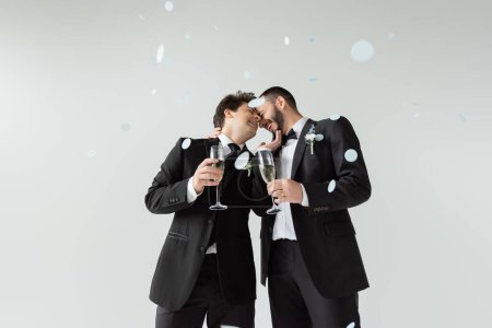 Joyeux mariés homosexuels en costumes se touchant tout en tenant des verres de champagne et en célébrant le mariage sous des confettis tombants sur fond gris