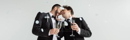 Unbekümmerte gleichgeschlechtliche Bräutigame in Anzügen, die sich berühren und Champagner halten, während sie ihre Hochzeit unter fallendem Festkonfetti auf grauem Hintergrund feiern, Banner 