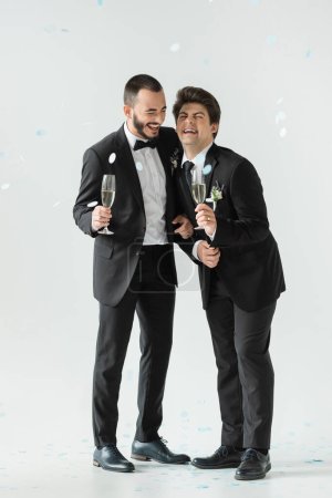 Full length of overjoyed same sex grooms in suits holding champagne and having fun under falling festive confetti during their wedding day on grey background