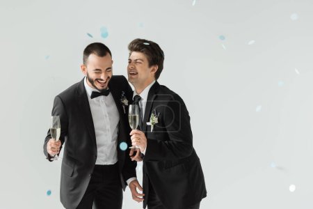 Cheerful gay groom holding champagne near elegant boyfriend in classic suit while standing under falling confetti during wedding on grey background
