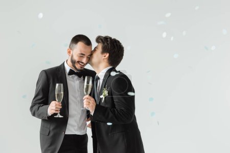 Young homosexual groom in formal wear kissing bearded boyfriend holding champagne under falling confetti on grey background
