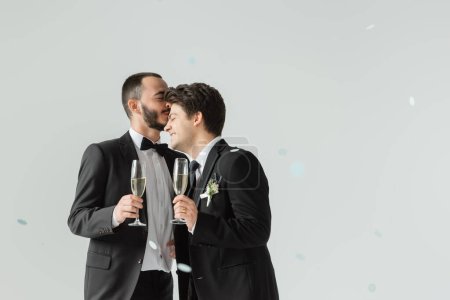 Bearded gay groom in formal wear holding glass of champagne and kissing smiling young boyfriend under confetti during wedding ceremony on grey background