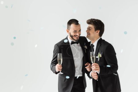Positive same sex grooms in classic attire holding champagne while standing under falling confetti during wedding ceremony on grey background