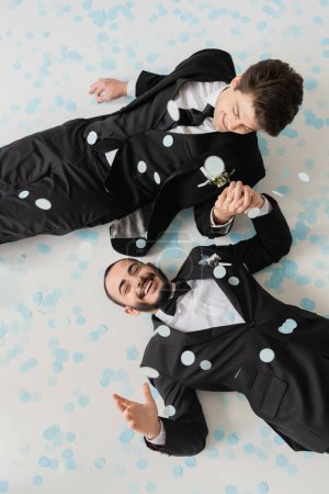 Foto de Vista superior de la alegre pareja del mismo sexo en trajes clásicos tomados de la mano mientras se divierten y tumbados en confeti durante la celebración de la boda sobre fondo gris - Imagen libre de derechos