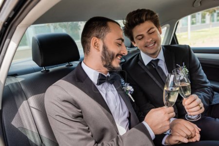 Smiling same sex grooms in formal wear toasting with glasses of champagne and holding hands while sitting on backseat of car after wedding, honeymoon trip 
