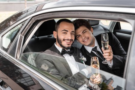 Smiling gay newlyweds in elegant classic attire with boutonnieres holding champagne and looking at camera from window while sitting on backseat of car during wedding trip 