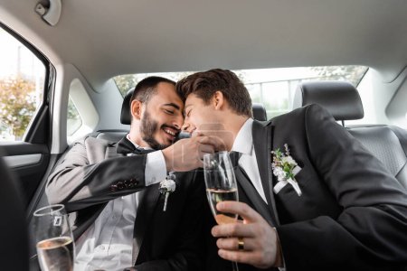 Carefree homosexual groom touching face of young boyfriend in braces and elegant suit with boutonniere and holding champagne while sitting on backseat of car