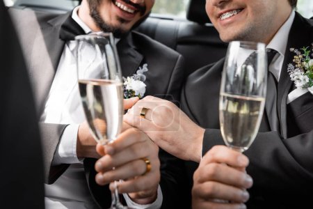 Ausgeschnittene Ansicht positiver gleichgeschlechtlicher Frischvermählter in klassischen Anzügen mit Boutonnieres an den Händen und verschwommenen Gläsern Champagner während einer Autofahrt 