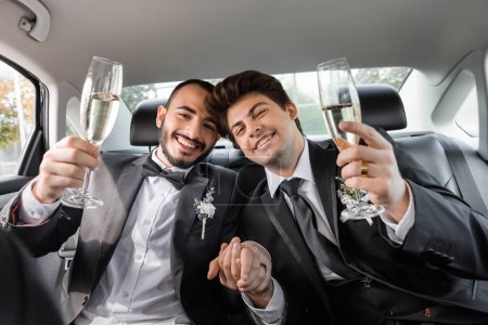 Smiling young gay newlyweds in formal wear holding hands and champagne while looking at camera during road trip while sitting on backseat of car 