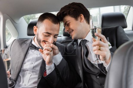 Positivo novio gay en elegante formal desgaste besos mano de joven pareja en frenillos y celebración de copa de champán durante viaje de luna de miel carretera y sentado en el asiento trasero del coche 