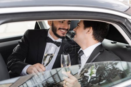 Positiver und bärtiger homosexueller Bräutigam im eleganten Anzug, der ein Glas Champagner in der Hand hält und seinen jungen Freund während der Flitterwochen auf dem Rücksitz des Autos ansieht 