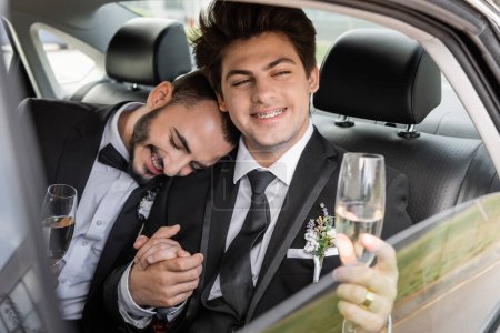Smiling and young gay groom with braces in elegant suit with boutonniere holding glass of champagne and hand of boyfriend while sitting on backseat of car during honeymoon 