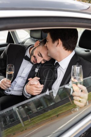 Smiling gay man in classic suit with boutonniere holding champagne and hand of bearded boyfriend with closed eyes while celebrating wedding in car during honeymoon 