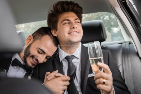 Carefree young gay man in formal wear and braces holding blurred glass of champagne and hand of bearded boyfriend after wedding celebration and sitting on backseat of car 