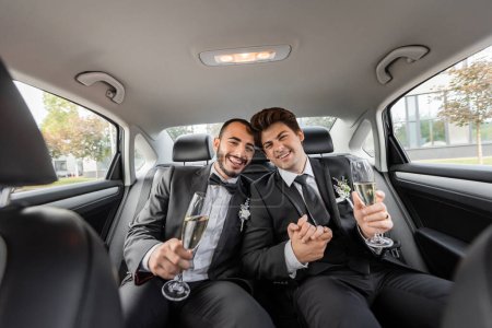 Smiling homosexual couple in classic suits with boutonnieres holding hands and glasses of champagne while celebrating wedding on backseat of car 
