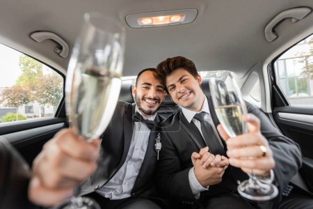 Cheerful homosexual grooms in classic suits holding blurred glasses of champagne and looking at camera during wedding celebration on backseat of car 