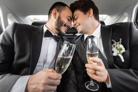 Lächelnde und junge homosexuelle Bräutigame in formeller Kleidung mit Boutonnieres sitzen Nase an Nase und halten Champagner auf dem Rücksitz des Autos nach der Hochzeitsfeier