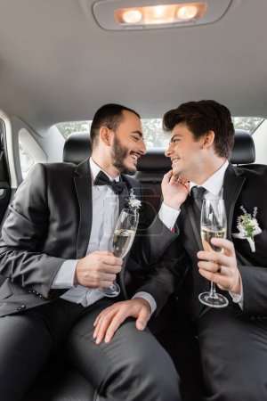 Téléchargez les photos : Vue latérale du marié gay souriant et barbu en costume classique touchant jeune petit ami dans des bretelles avec verre de champagne tout en célébrant le mariage sur le siège arrière de la voiture pendant la lune de miel - en image libre de droit