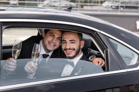 Lächelnde gleichgeschlechtliche Bräutigame in offizieller Kleidung halten Champagnergläser in der Hand und blicken durch das Fenster in die Kamera, während sie auf dem Rücksitz des Autos sitzen, bevor sie in die Flitterwochen fahren 