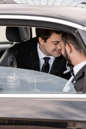 Smiling same sex grooms in classic suits holding champagne glasses and kissing after wedding celebration on backseat of car before honeymoon trip 