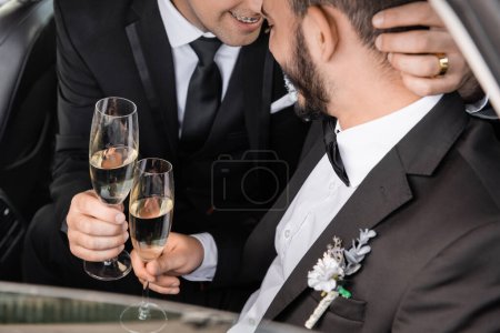 Smiling homosexual groom in formal wear holding champagne glass and hugging bearded boyfriend while celebrating wedding in car during honeymoon 
